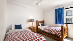 A bedroom with two single beds, a nightstand, a lamp, and a window with blue curtains. - Phoenix 8 Coolum Beach (Coolum Beach)