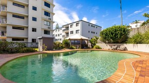 A swimming pool with a brick border, surrounded by a garden with plants and a wooden fence. - Phoenix 8 Coolum Beach (Coolum Beach)