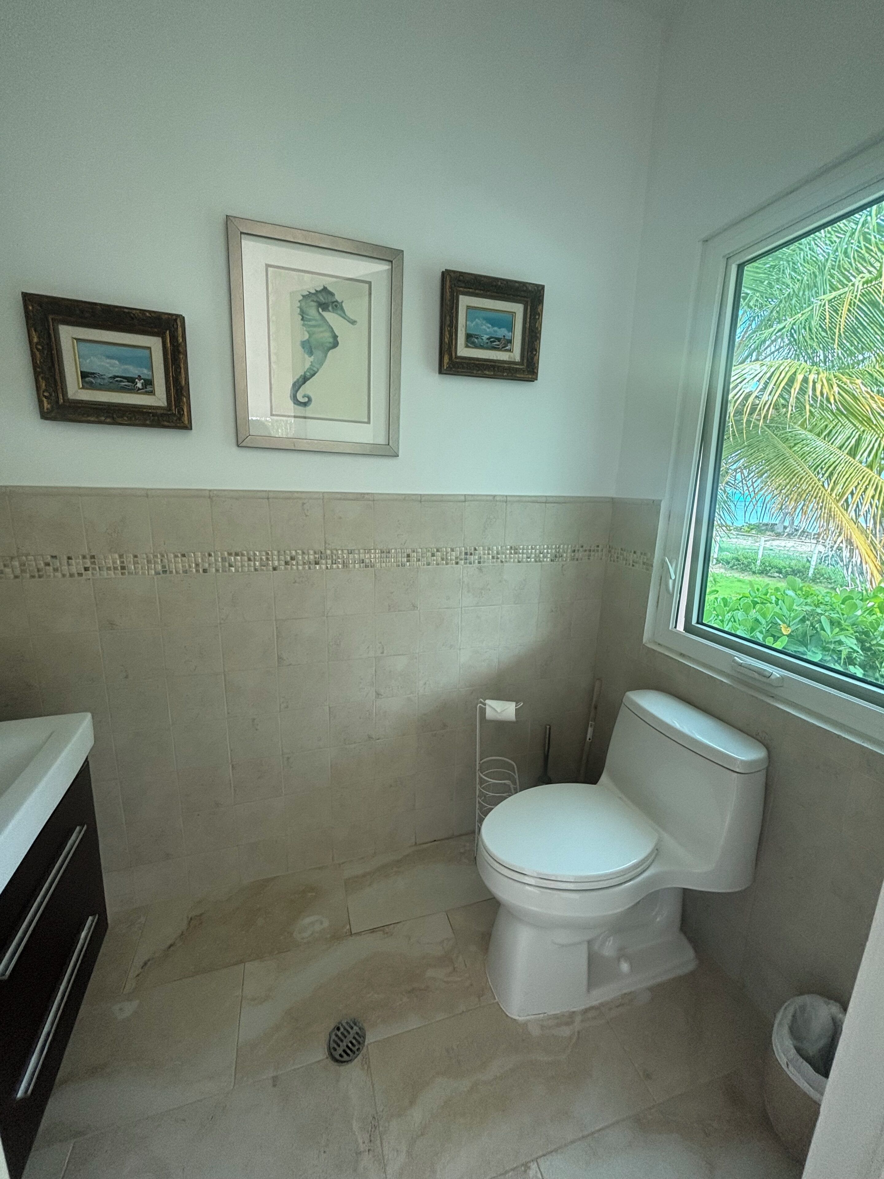 A small bathroom with a toilet, a sink, and a window. There are three framed pictures on the wall.