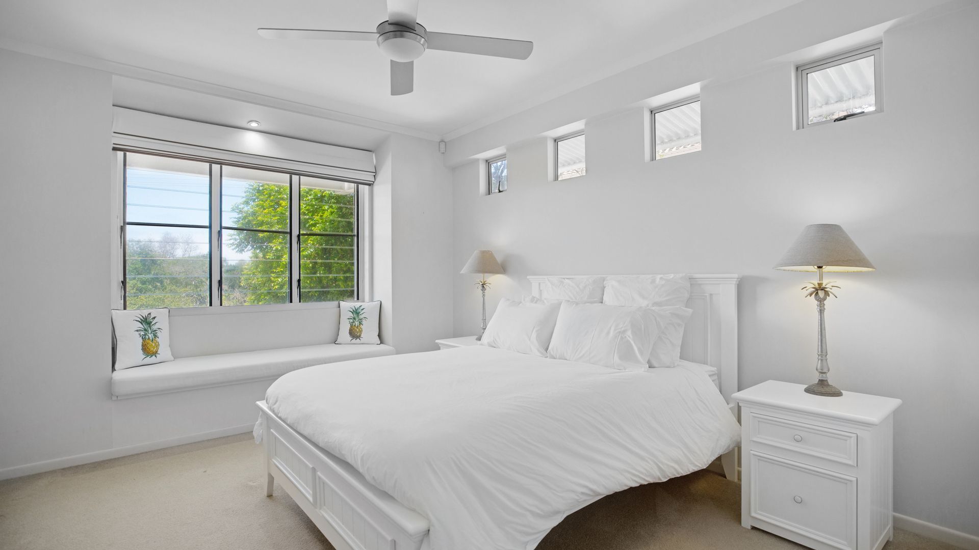 A bedroom with a bed, nightstands, a ceiling fan, and a window with a view of greenery.