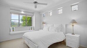 Ein Schlafzimmer mit Bett, Nachttischen, Deckenventilator und Fenster mit Blick auf Grünflächen.