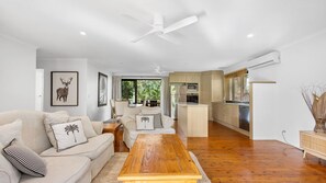 A modern living room with a sofa, coffee table, and artwork, leading to a kitchen with wooden cabinets and a dining area. - Gannet's Nest Peregian Beach (Peregian Beach)