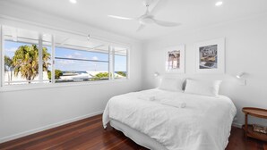 Una camera da letto con un letto grande, una ventola a soffitto e vista su palme e spiaggia dalla finestra.
