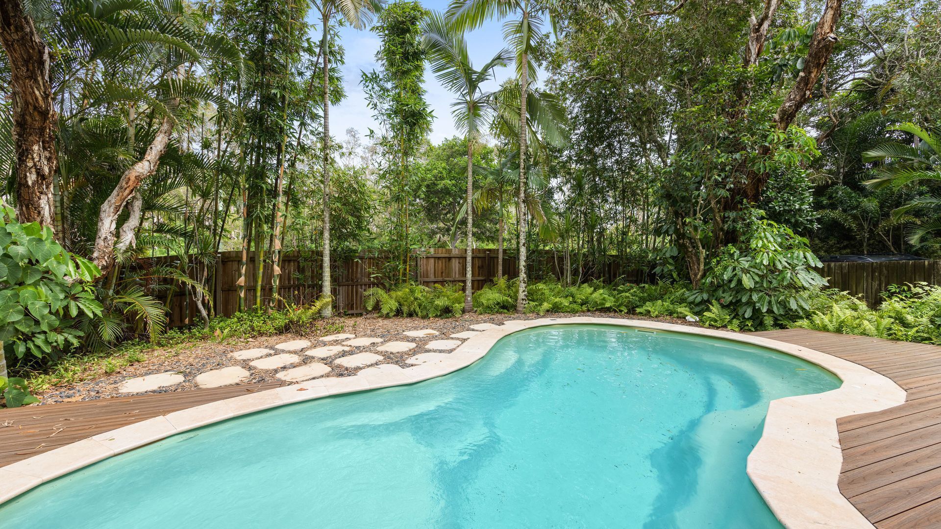 A swimming pool with a wooden deck, surrounded by lush tropical vegetation and a wooden fence.