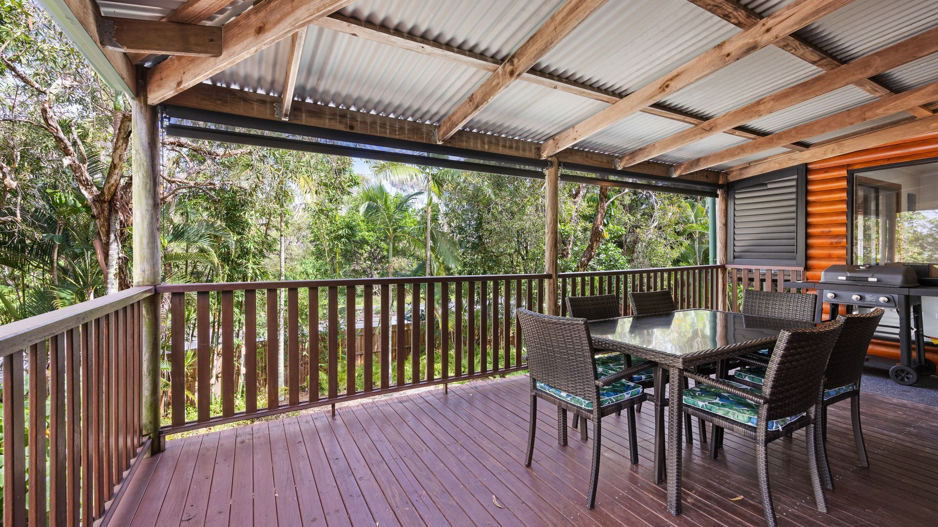 Una terrazza esterna riparata con tavolo in vetro e sedie in vimini, circondata da ringhiera in legno e travi metalliche.