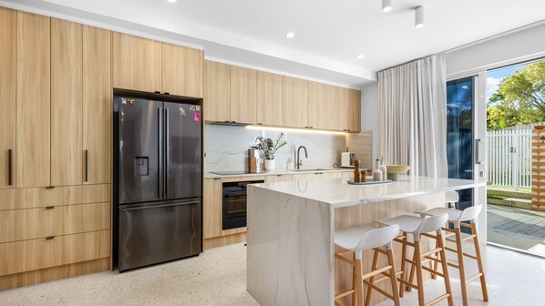 A modern kitchen with a stainless steel refrigerator, wooden cabinets, a marble countertop, and a bar area with stools. - Olas by The Sea 2 Peregian Beach (Peregian Beach)