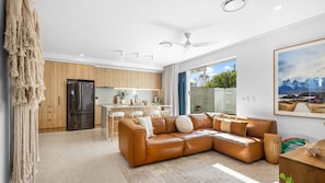A modern living room with a leather sectional sofa, a kitchen with wooden cabinets, and a large window with a view of greenery. - Olas by The Sea 2 Peregian Beach (Peregian Beach)