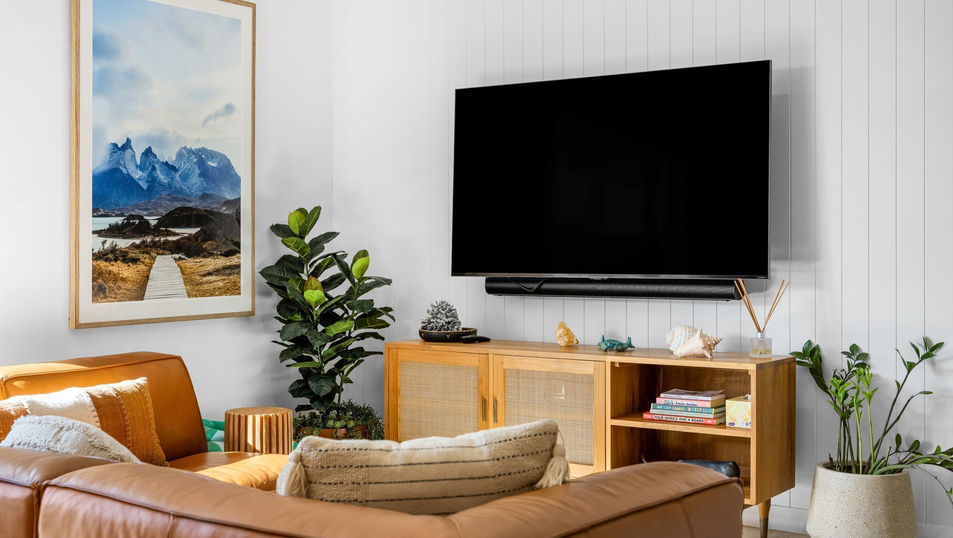 Un salon doté d’un téléviseur à écran plat fixé au mur, d’un canapé, d’un meuble en bois orné d’objets décoratifs et d’un grand tableau encadré représentant un paysage de montagne.