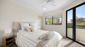 A bedroom with a bed, a nightstand, a ceiling fan, a window with a view, and a balcony. - First Avenue Glade 6 (Coolum Beach)