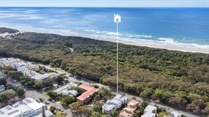 An aerial view of a coastal area with a dense forest, a beach, and residential buildings. - First Avenue Glade 6 (Coolum Beach)