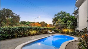 A swimming pool with lounge chairs, surrounded by a hedge and trees, with a building in the background. - First Avenue Glade 6 (Coolum Beach)
