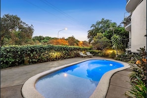 A swimming pool with lounge chairs, surrounded by a hedge and trees, with a building in the background. - First Avenue Glade 6 (Coolum Beach)