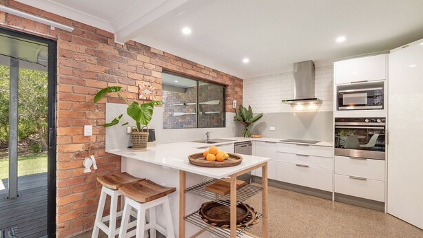 A modern kitchen with white cabinets, a brick accent wall, a large window, and built-in ovens. - Ashton Villa 1 Peregian Beach (Peregian Beach)