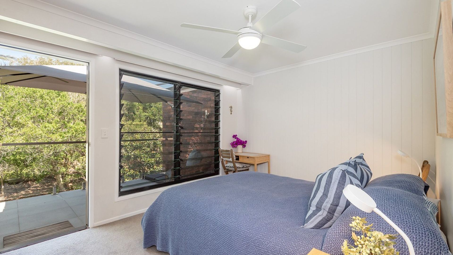 Une chambre avec une grande porte-fenêtre coulissante donnant sur un balcon, un ventilateur de plafond et un lit avec une literie bleue.