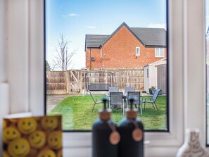 Blick durch ein Fenster auf ein Backsteinhaus mit einem umzäunten Garten, Gartenmöbeln und blauem Himmel.