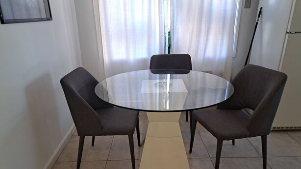 Una taula rodona de vidre amb quatre cadires fosques a una habitació amb parets blanques i un sòl de rajola.