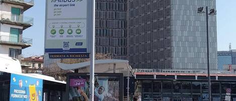 A bus stop with a sign for Alibus, a tall building in the background, and a bus with advertisements.