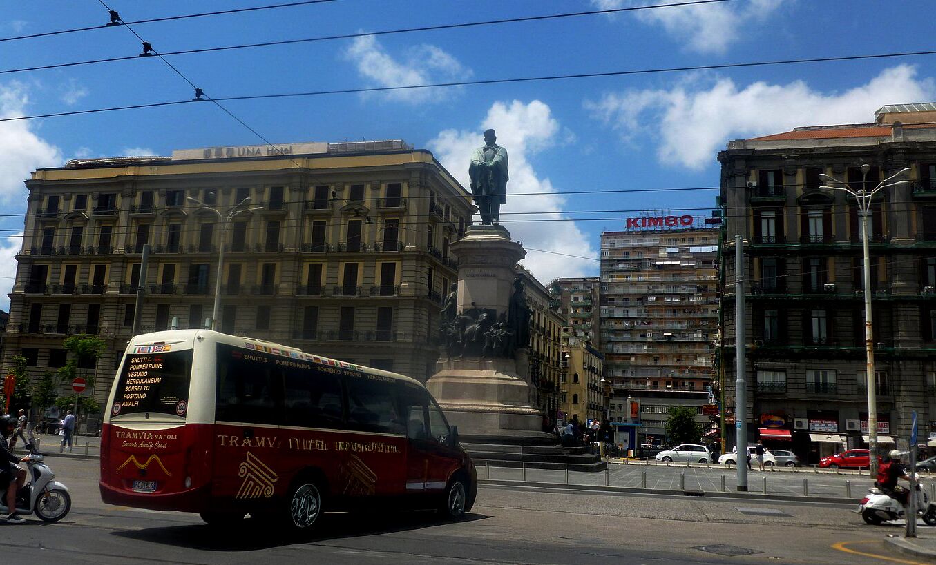 A red and white bus with "TRAMVIA NAPOLI" on the side, driving on a city street with buildings and a statue in the background.