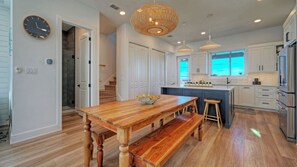 A modern kitchen with a wooden dining table, built-in appliances, and a large window. - Dune & Done by Avantstay Outdoor Lounge + Firepit Shared Pool + Golf Cart Zone Mustang Island Beach (Port Aransas)