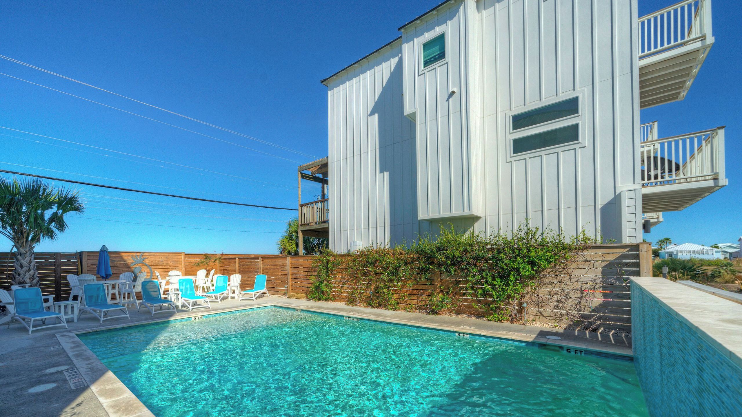 A swimming pool with lounge chairs and a wooden deck, a white building with balconies, and a clear blue sky.