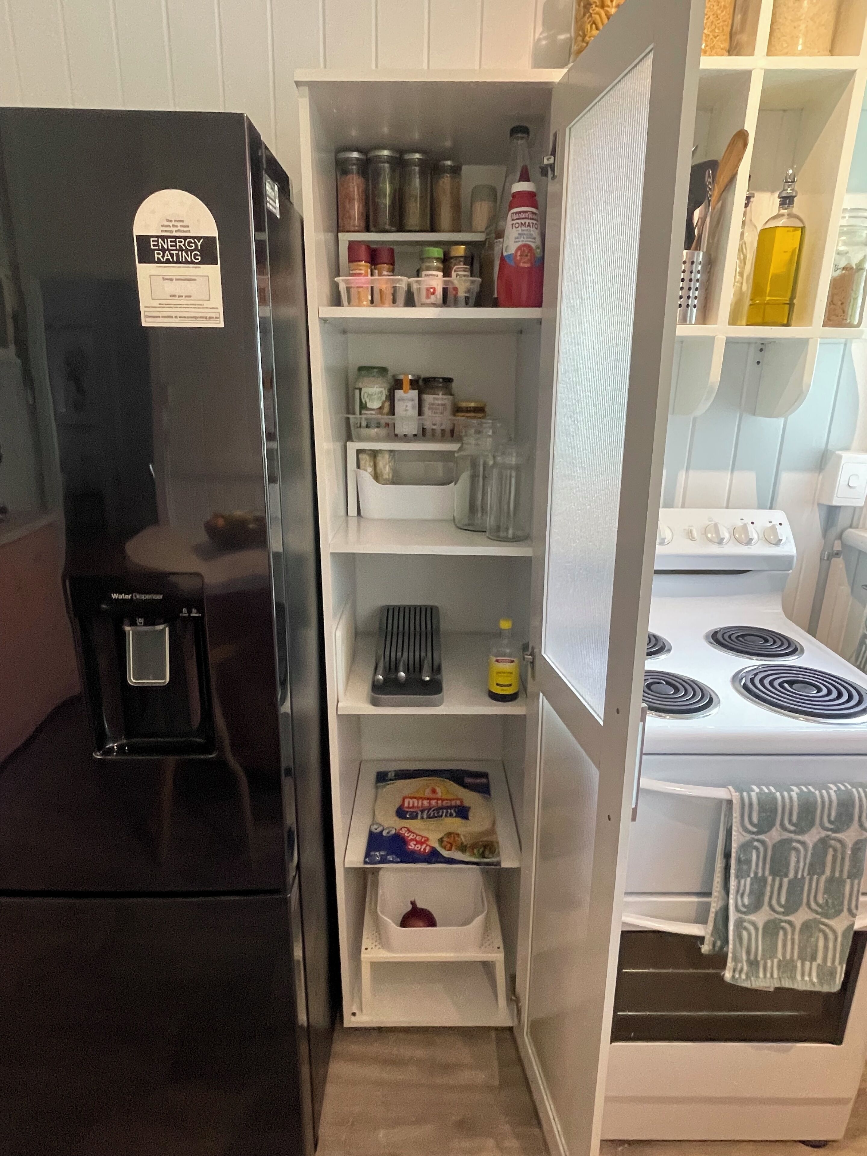 Una cocina compacta con un refrigerador negro, un dispensador de agua, una estufa y repisas repletas de frascos y artículos esenciales de cocina.