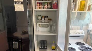 Una cocina compacta con un refrigerador negro, un dispensador de agua, una estufa y repisas repletas de frascos y artículos esenciales de cocina.