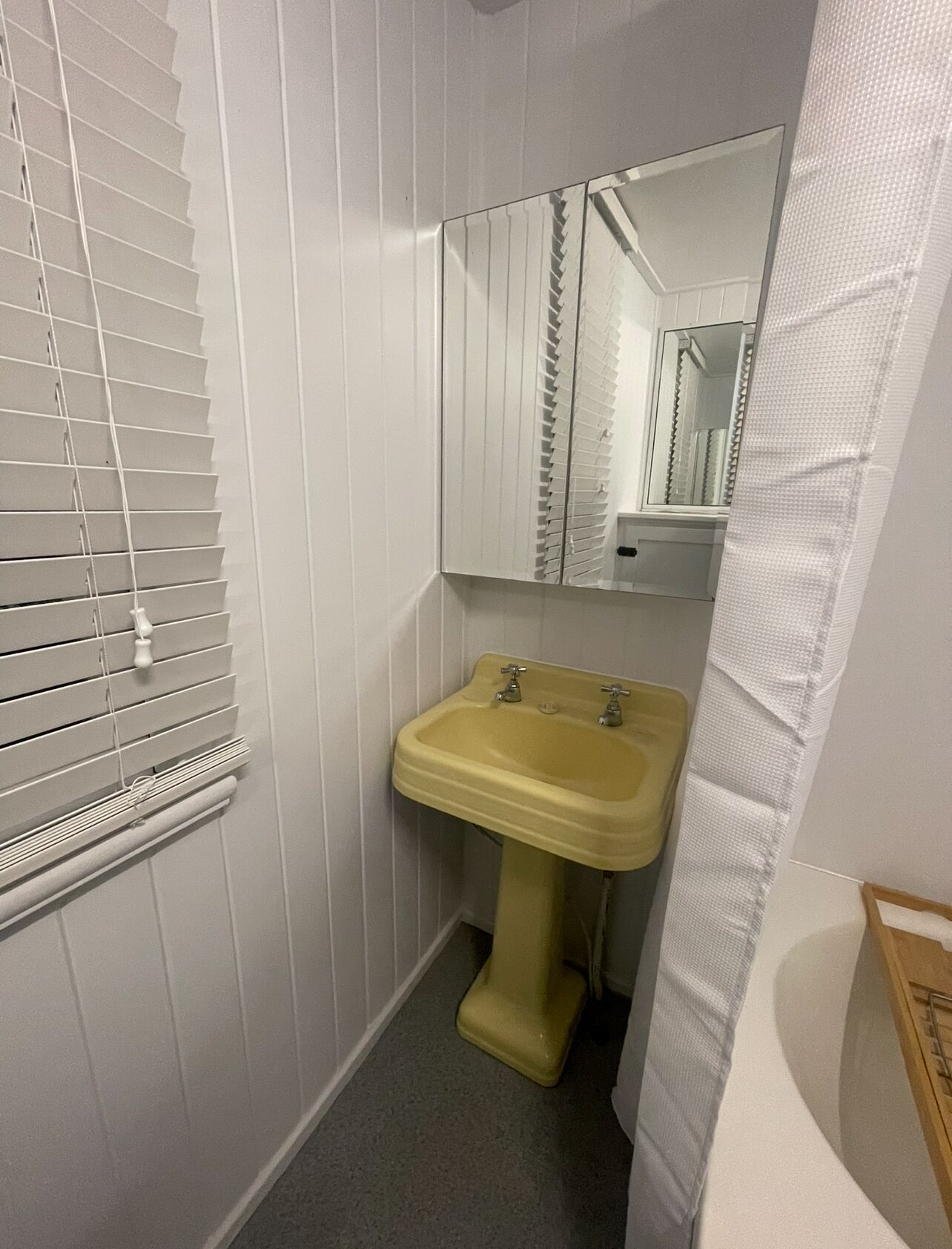Un baño estrecho con un lavamanos amarillo, un espejo y una ventana con persianas.