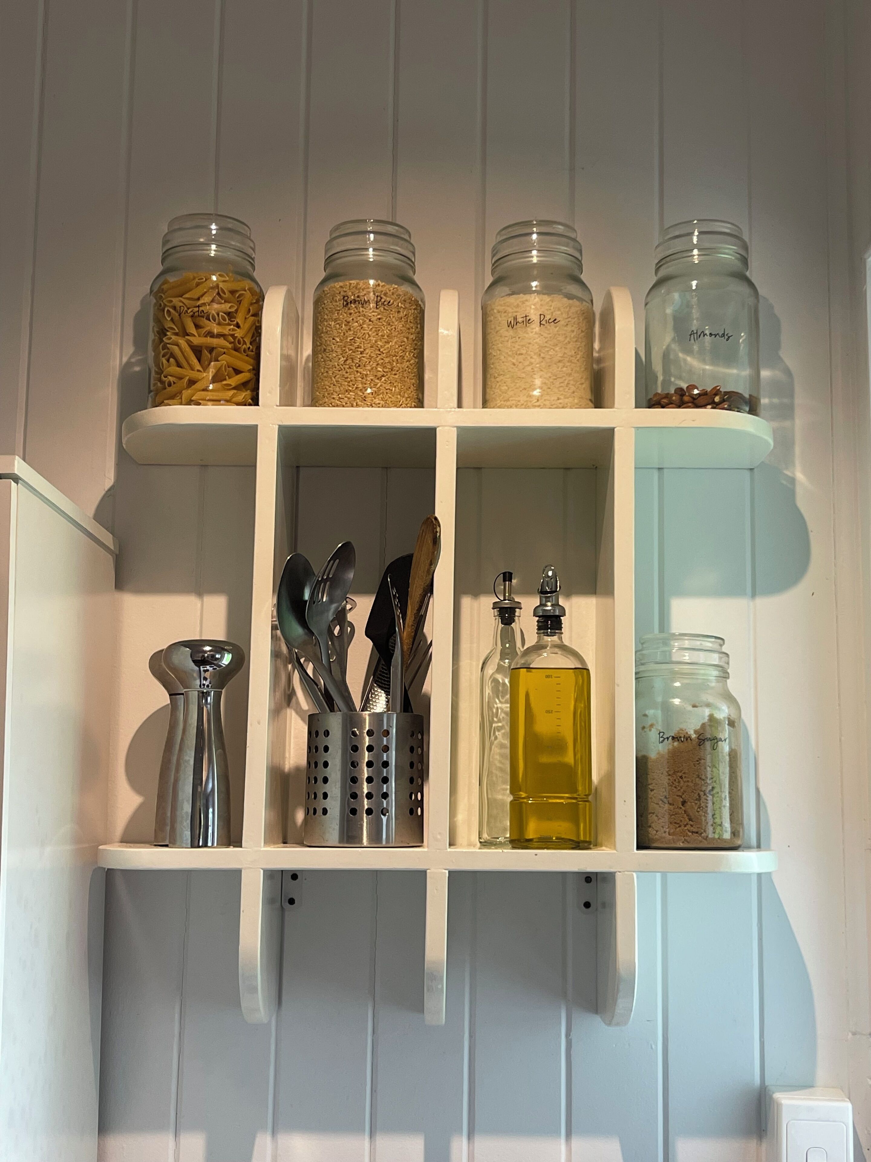 Une étagère de cuisine contenant des bocaux étiquetés « pâtes », « riz brun », « riz blanc », « amandes » et « sucre brun ».