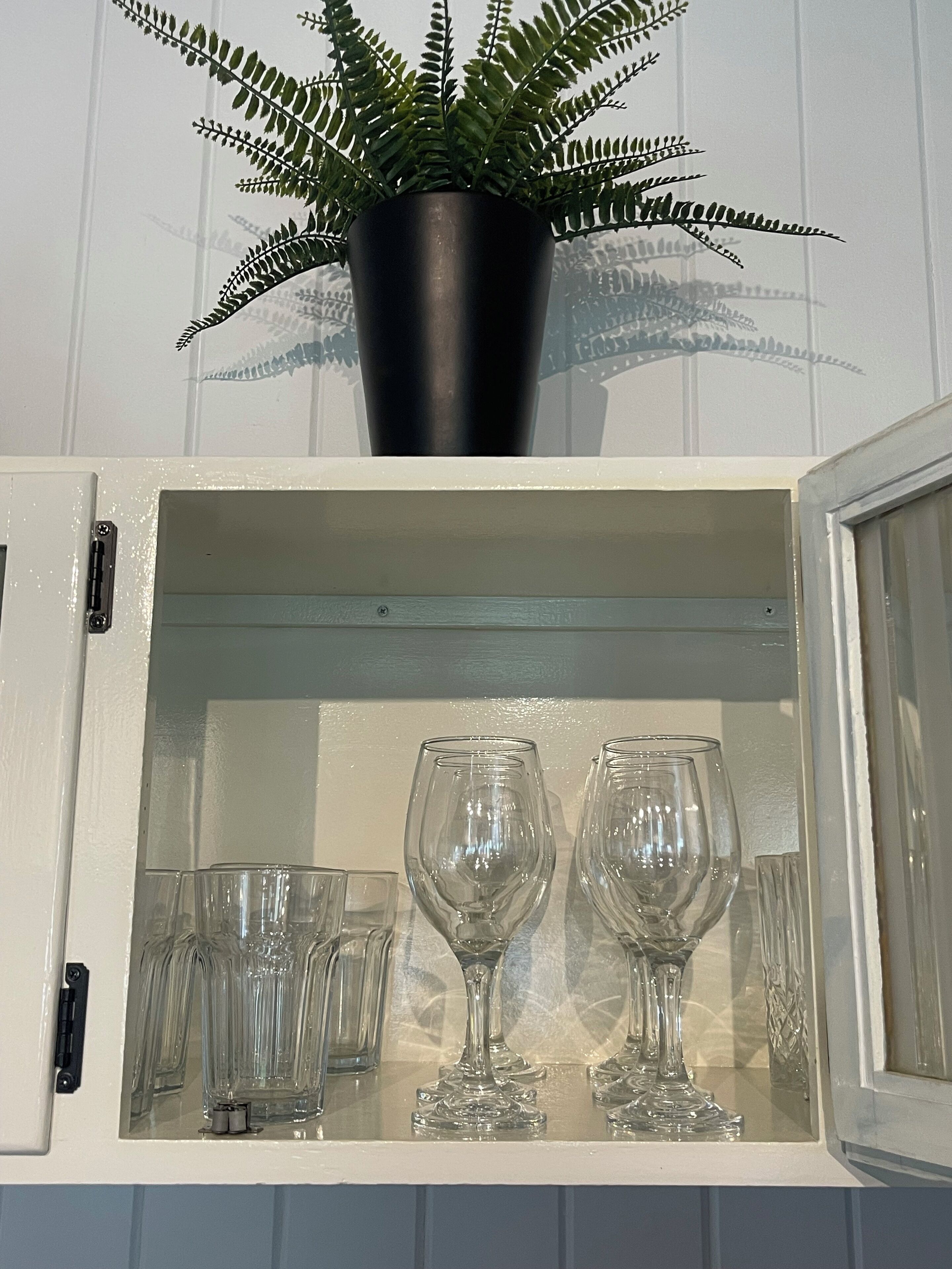 A cabinet with a potted fern on top and several wine glasses inside.