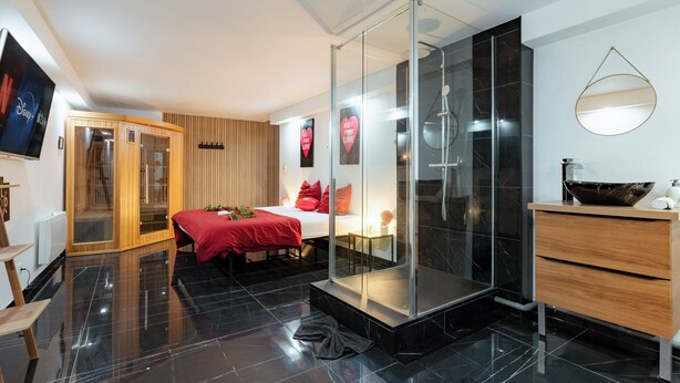 A modern hotel room with a glass-enclosed shower, a wooden wardrobe, a bed with red bedding, and a flat-screen TV mounted on the wall.
