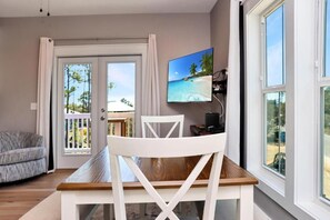 Ein Zimmer mit Esstisch aus Holz, weißen Stühlen, einem an der Wand montierten Fernseher und Blick auf einen Strand durch Glastüren.