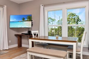 Ein Zimmer mit Holztisch, weißen Stühlen, einem Fernseher an der Wand und einem großen Fenster mit Blick auf Bäume und einen Strand.