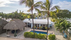 Ein Resort mit Pool, strohgedeckten Gebäuden, Palmen und einem weißen Gebäude mit Metallbedachung.