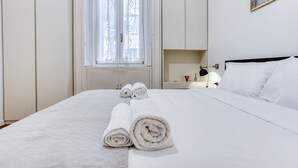 Ein ordentlich bezogenes Bett, gerollte Handtücher, ein Fenster mit Vorhängen, ein Nachttisch mit Lampe und eine Karte an der Wand.