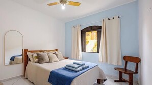 Un dormitorio con cama, ventilador de techo, ventana con cortinas, una silla y un espejo en la pared.