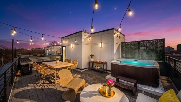 A rooftop patio with a hot tub, outdoor seating, string lights, and a city view at dusk. - 2 Homes | 24 Guests | Rooftop Hot Tub Fire Pit (Nashville)