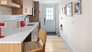 Una cocina con armarios de madera, encimera blanca, hervidor rojo y una silla de mimbre.