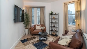 A modern living room with a leather sofa, a round coffee table, a bookshelf, and a large window with curtains. - Charming 2-Bed flat Steps from the Royal Mile - Pass the Keys (Edinburgh)