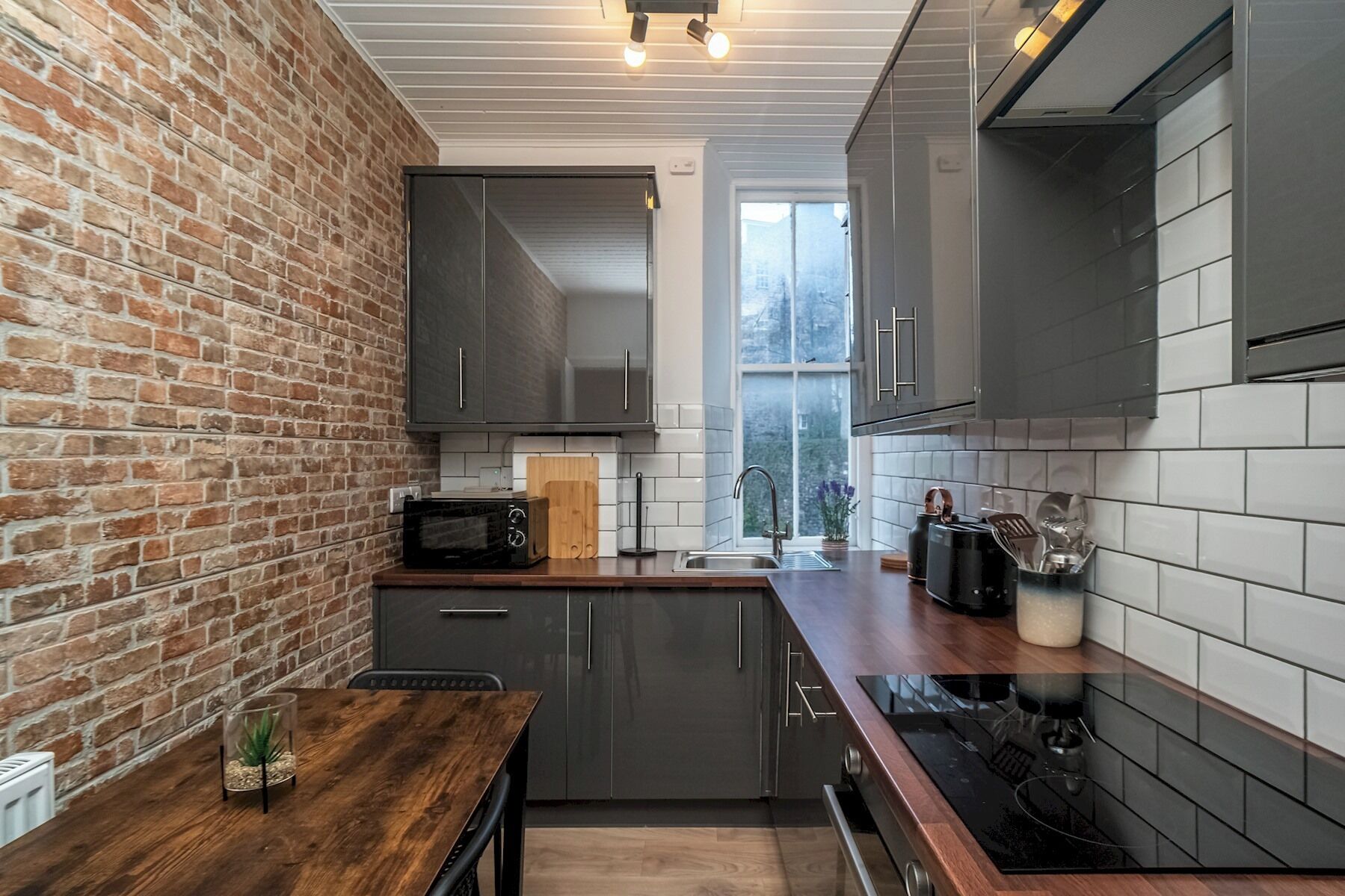 A modern kitchen with a brick wall, grey cabinets, a wooden countertop, and a window providing natural light.