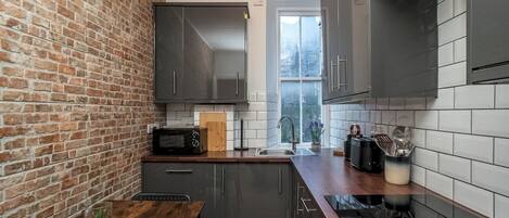 A modern kitchen with a brick wall, grey cabinets, a wooden countertop, and a window providing natural light.