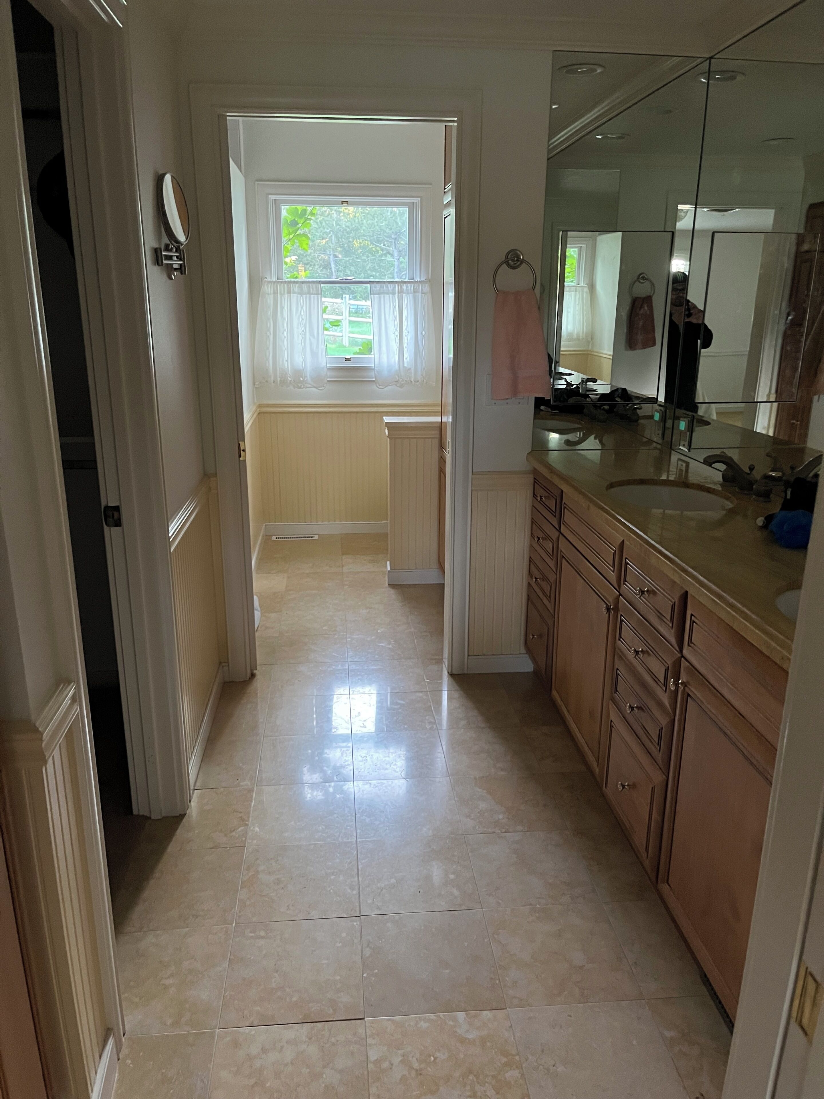 A narrow hallway leading to a bathroom with a large mirror, a sink, and a window with curtains.