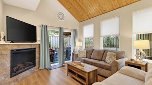 A living room with a fireplace, a sofa, a television, and a view of the outdoors. - Tides & Timber by Avantstay Lakeside Retreat + Hot Tub + Kayaks + Paddleboard + Bikes Near Beach (Rockaway Beach)