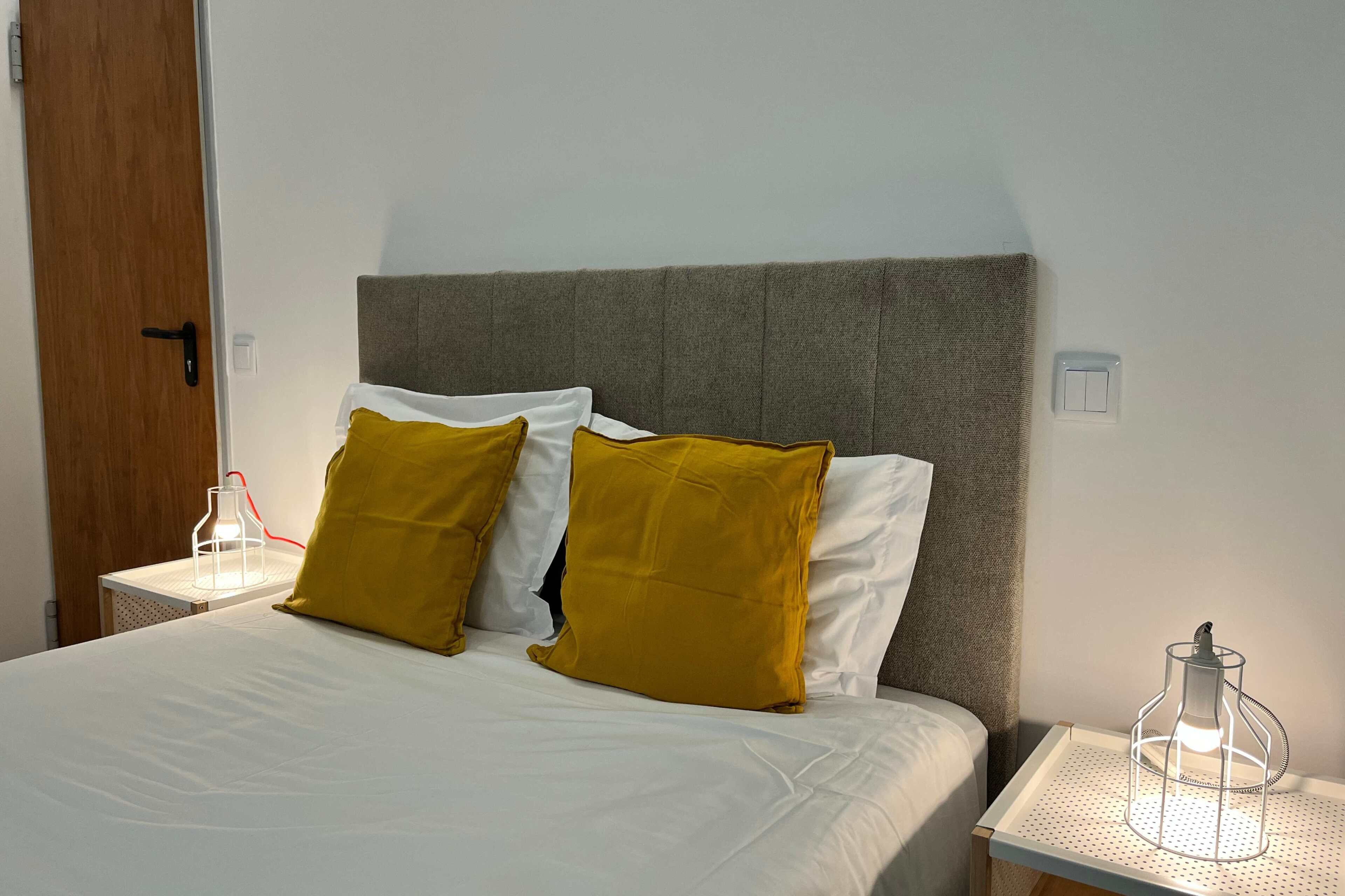 A neatly made bed with white and mustard pillows, a grey headboard, and a bedside table with a lamp and a small decorative item.