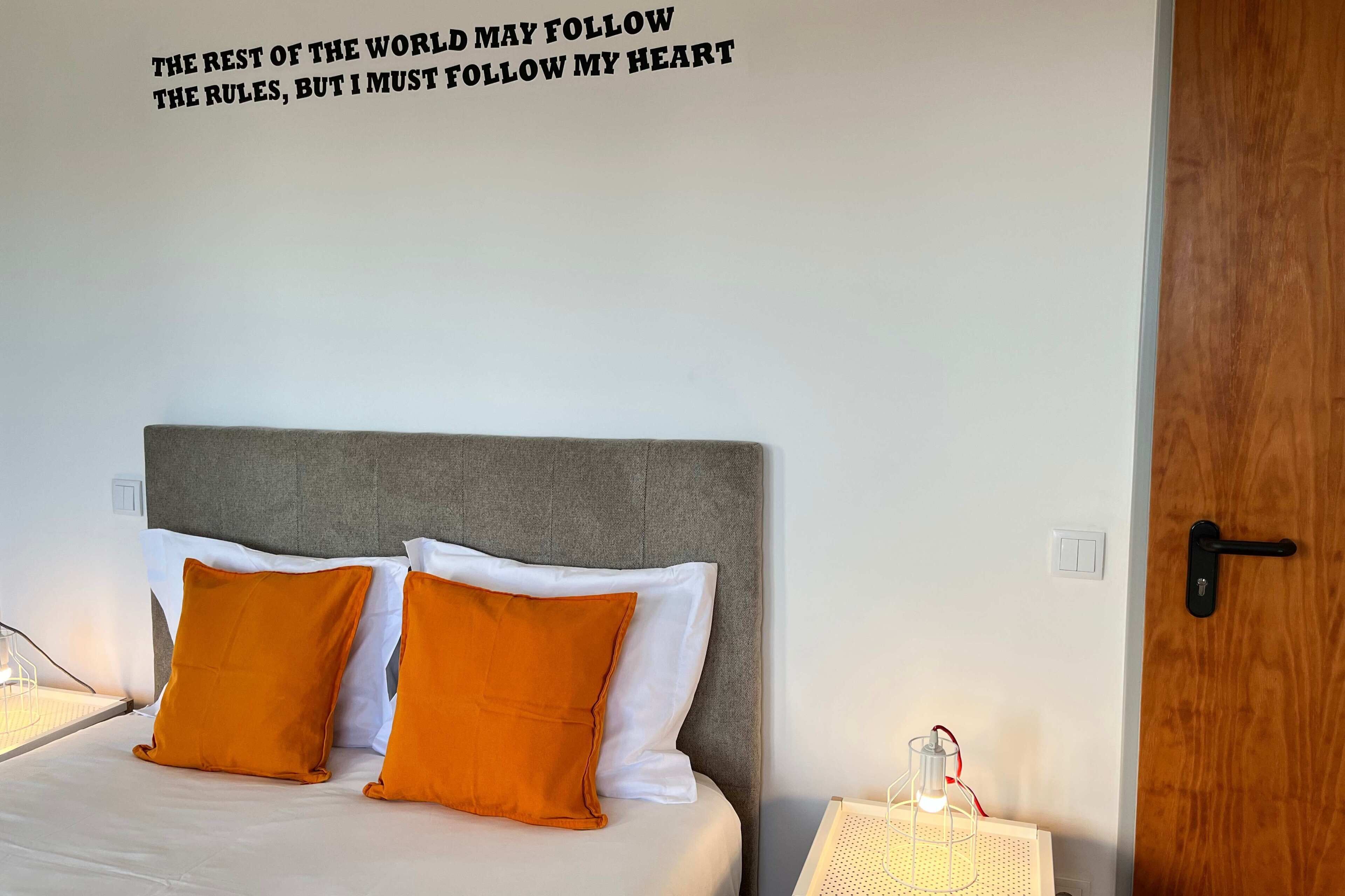 A neatly made bed with white and orange pillows, a grey headboard, and a motivational quote on the wall.