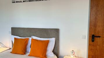 Un lit bien fait, avec des oreillers blancs et orange, une tête de lit grise et une citation inspirante accrochée au mur.