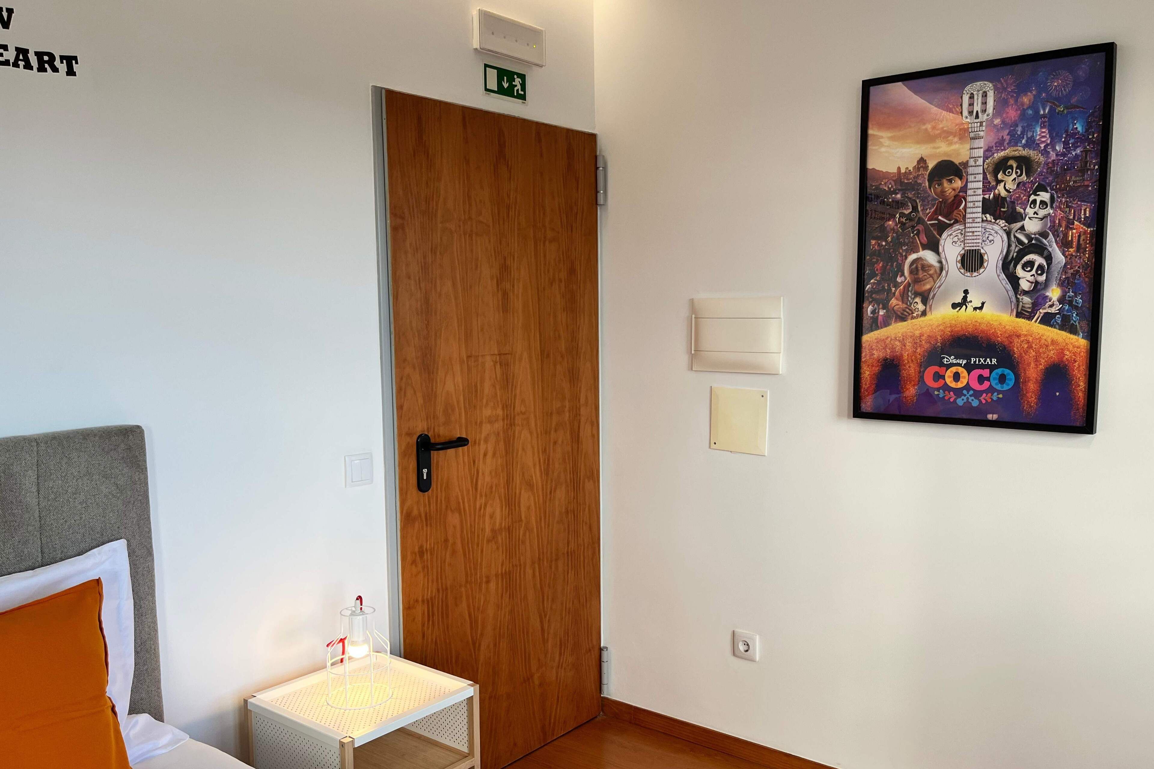 Una habitación de hotel con una puerta de madera, una pequeña mesa con una lámpara, una cama con un cojín naranja y un cartel de 'Coco' en la pared.