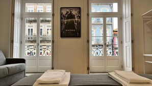 Habitación de hotel con cama, sofá gris, un cartel en la pared y vistas de edificios a través de las puertas.