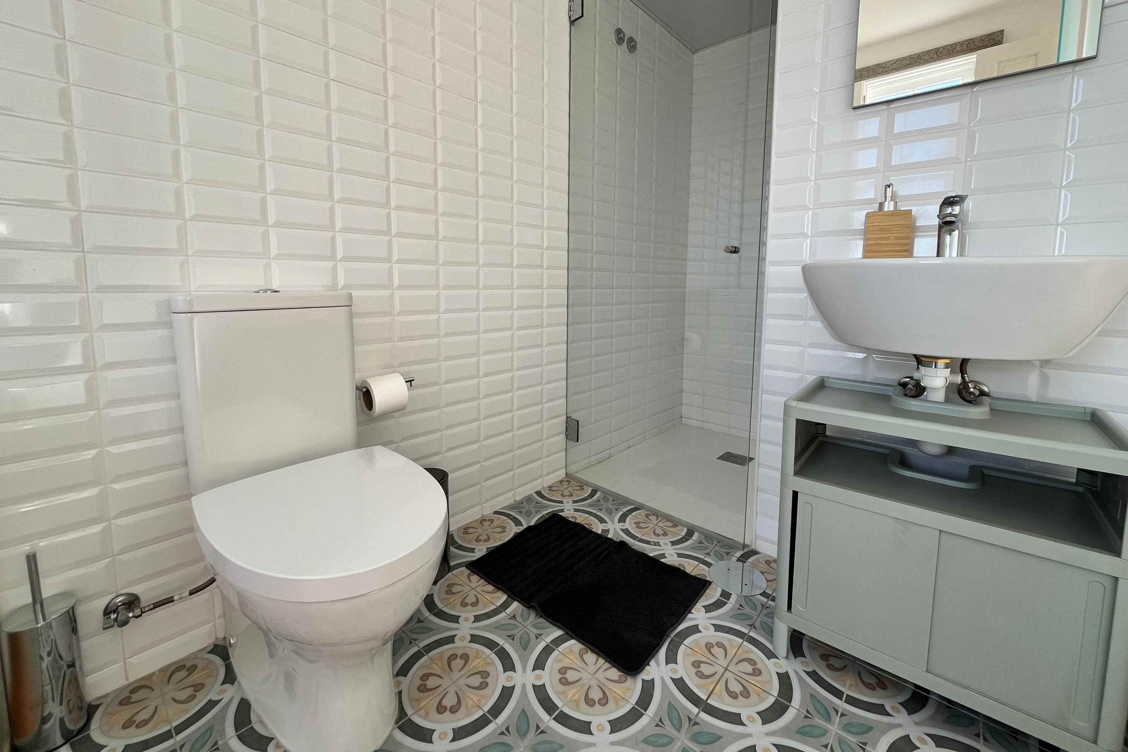 A bathroom with white tiled walls, a white toilet, patterned floor tiles, a black bath mat, and a white sink with a wooden soap dispenser.