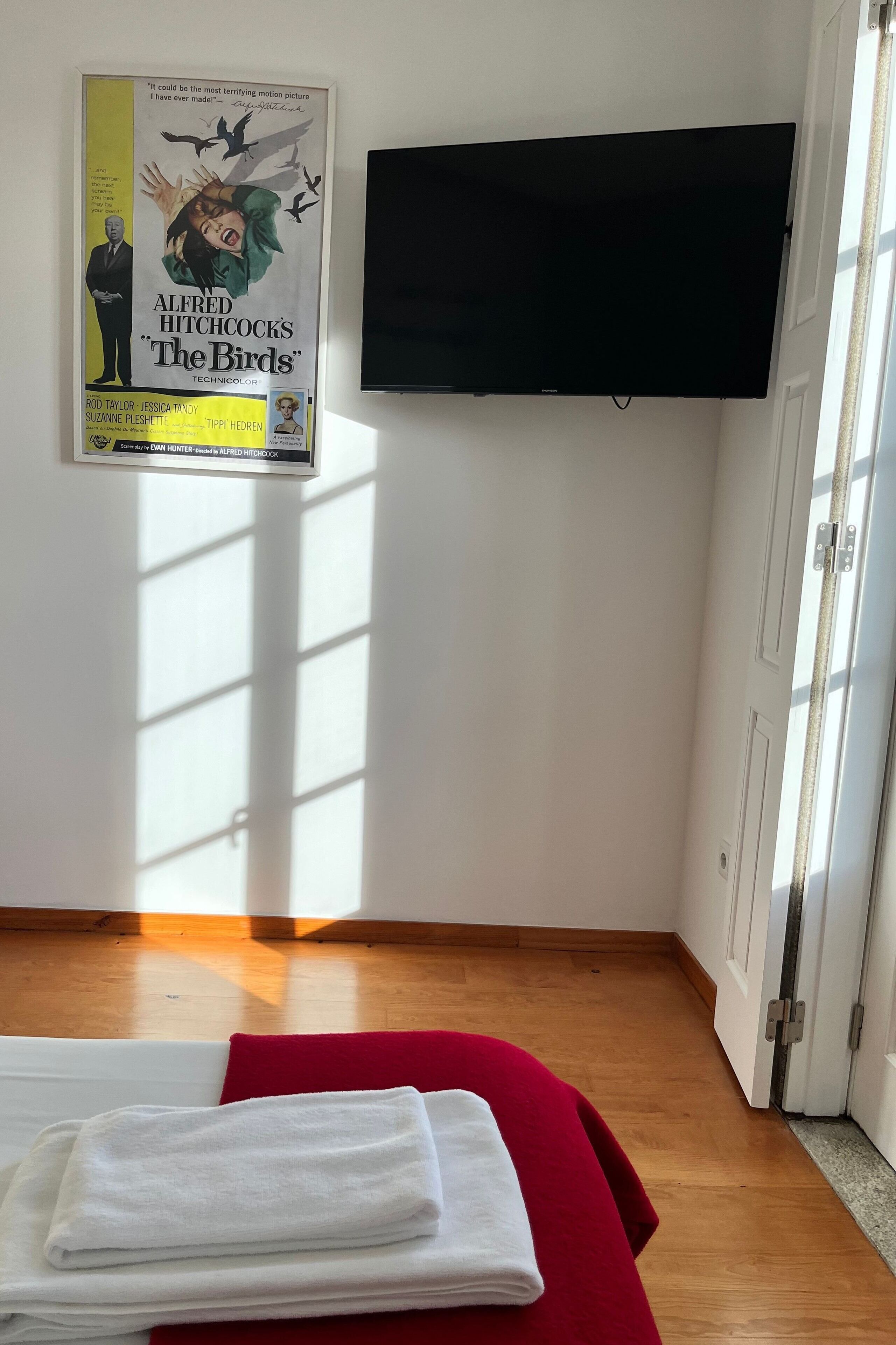 Une chambre avec un lit, une affiche du film « Les Oiseaux » d’Alfred Hitchcock et un téléviseur à écran plat fixé au mur.