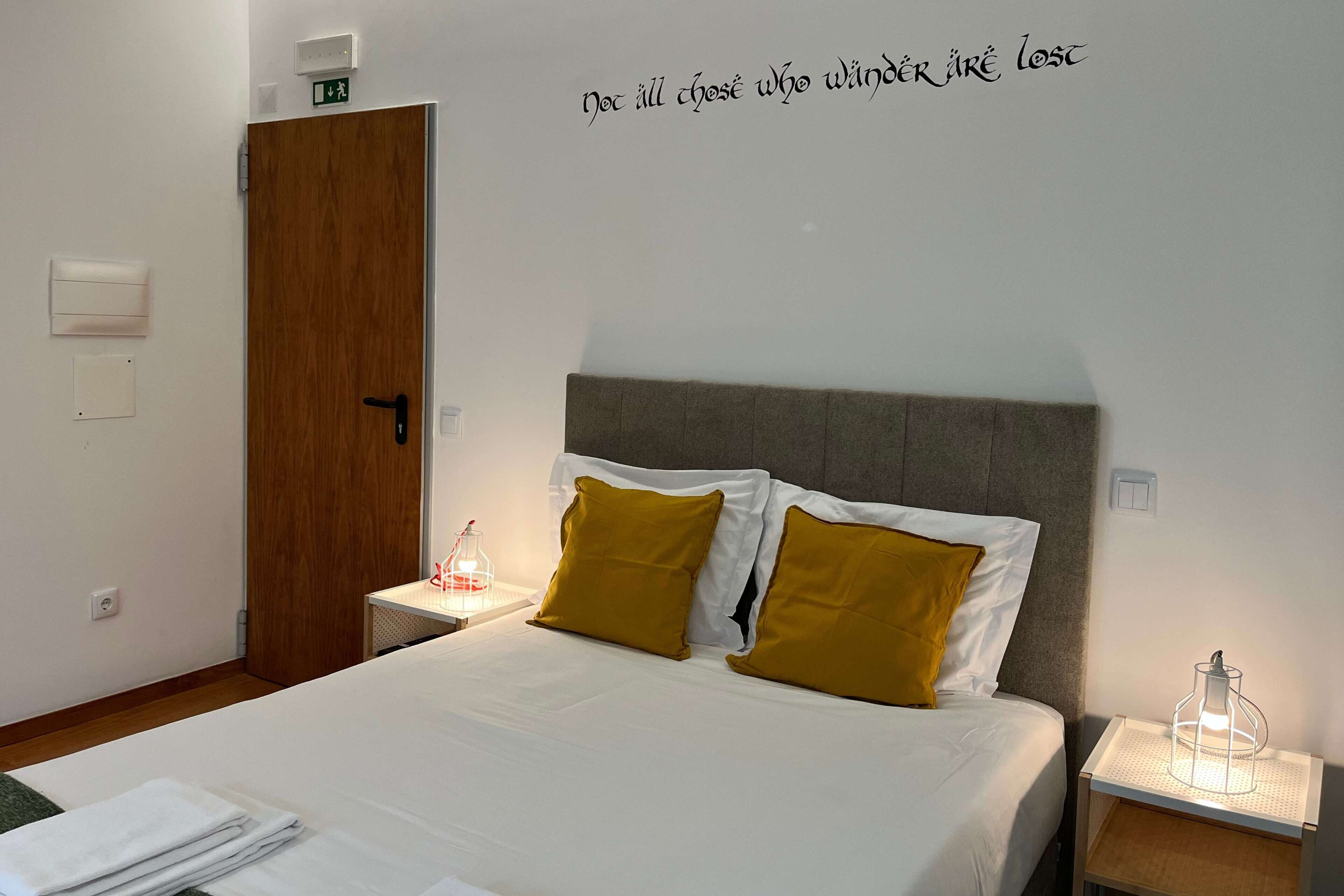 A neatly made bed with white linens and two mustard-colored pillows, a wooden door, and a wall-mounted light fixture.
